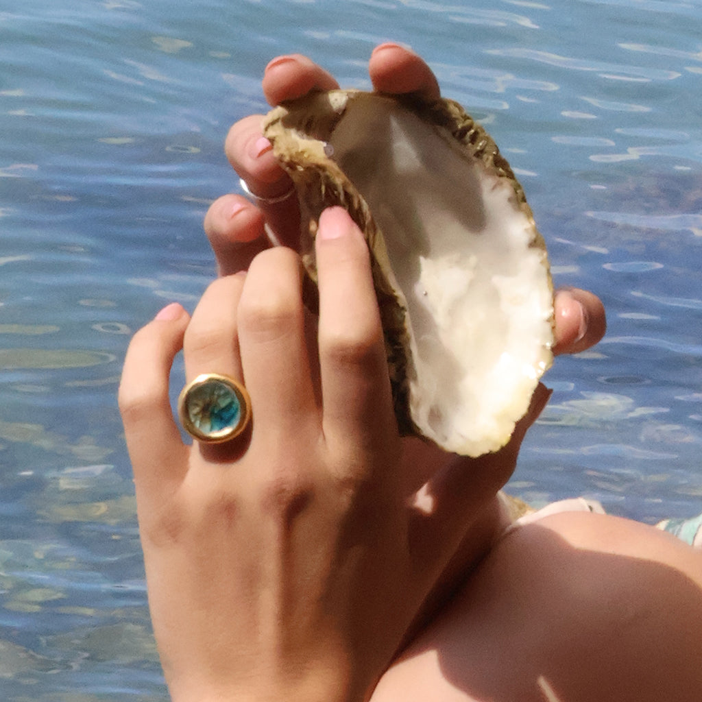 A female's hands hold an oyster shell in front of an ocean background. On her left hand, she wears a large round ring made from ceramic and teal, white and brown glass with gold detailing. 