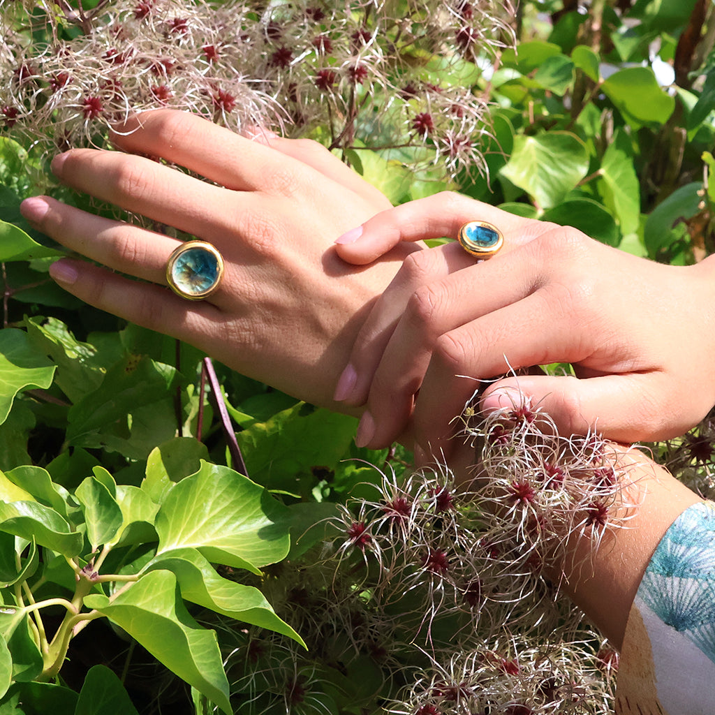 Two female hands wearing sterling silver rings with round ceramic, glass and gold charms. The glass is teal, white and brown and the hands rest on top of green and pink foliage. 
