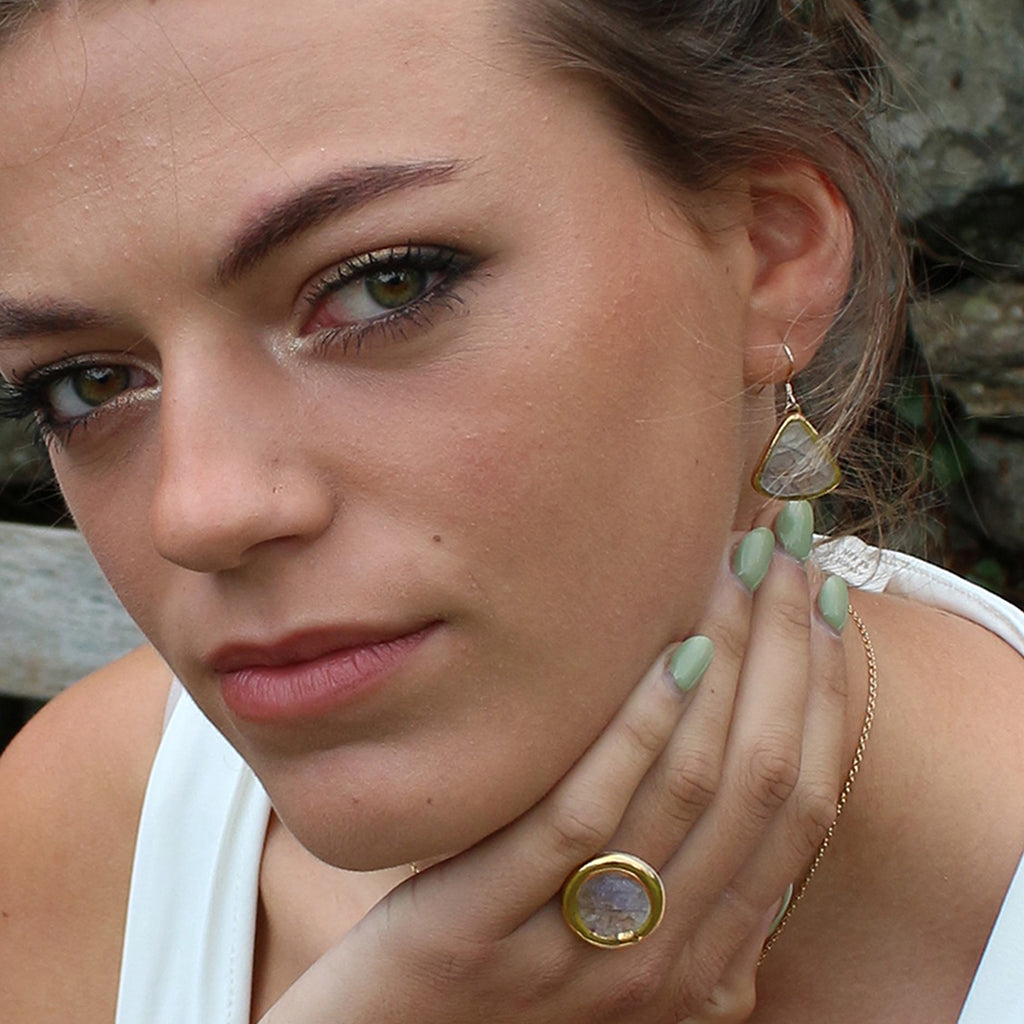 Close-up of a woman wearing gold earrings and a ring, with a blurred natural background