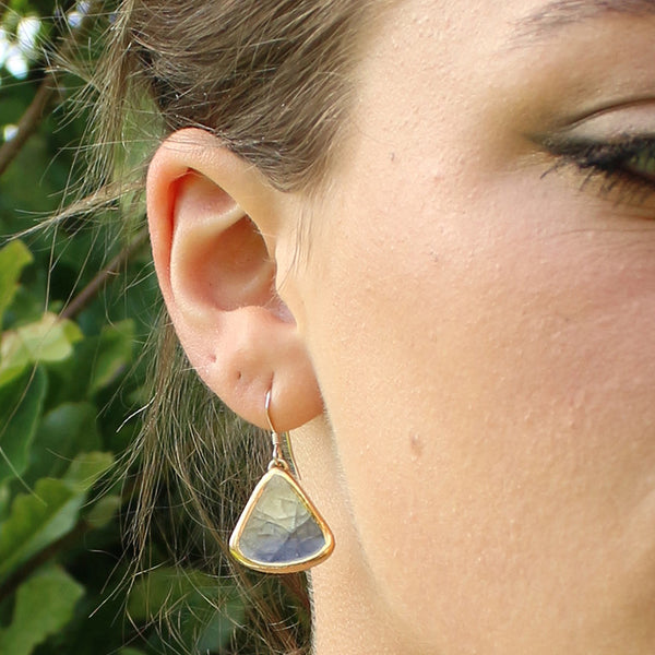 Close-up of a person wearing a gold earring with a blue gemstone against a blurred green background