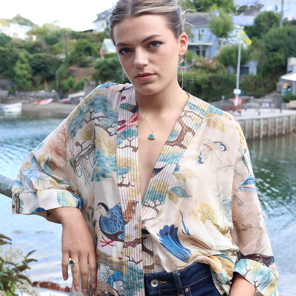 A woman standing outdoors by the water with a harbor and houses in the background. She wears teal-coloured jewellery pieces made from ceramic, glass and gold. 