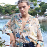 A woman standing outdoors by the water with a harbor and houses in the background. She wears teal-coloured jewellery pieces made from ceramic, glass and gold. 