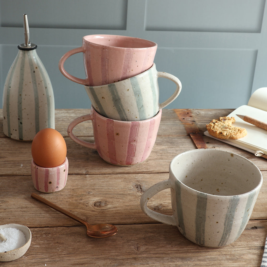 A set of handmade ceramic mugs in a pink stripe and a blue and white stripe design, styled on a rustic wooden table with an egg in an pink striped egg cup, a blue and white stripe mug and oil pourer. 
