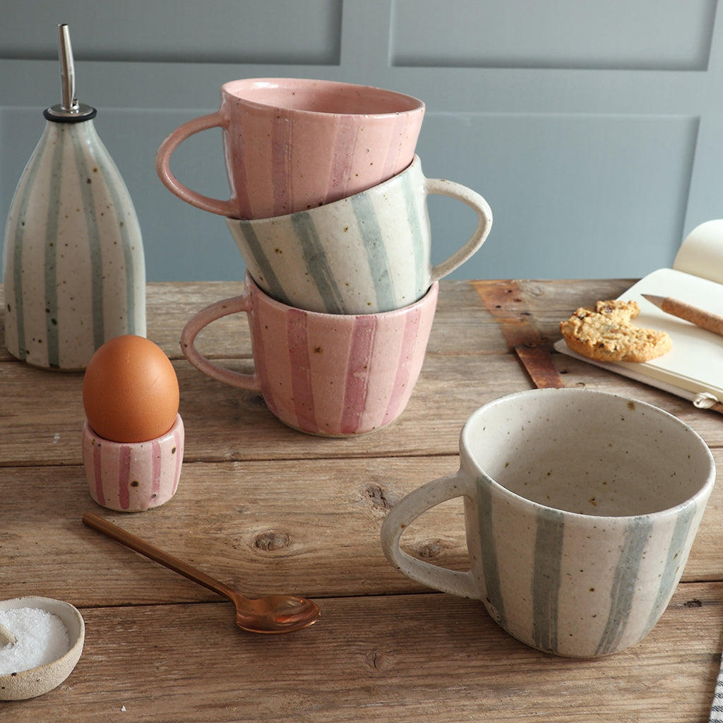 A set of handmade speckled mugs in pink, white, and blue stripes, styled on a rustic wooden table with a matching striped egg cup and oil pourer, a spoon, and an open notebook.