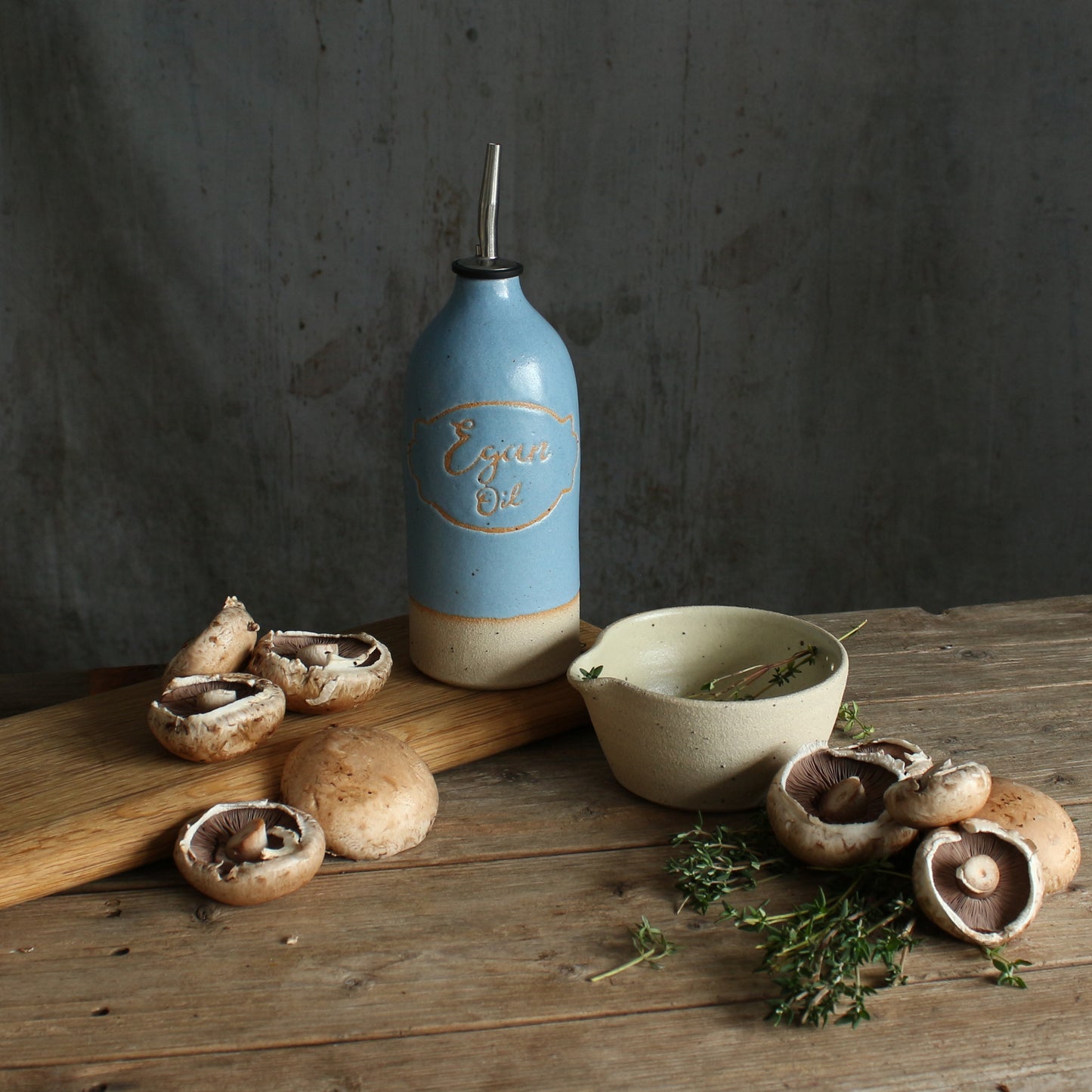 A rustic kitchen scene with a blue ceramic personalised oil pourer on a wooden table, surrounded by mushrooms, our handmade stoneware herb bowl bowl, and herbs against a dark gray wall.