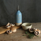 A rustic kitchen scene with a blue ceramic personalised oil pourer on a wooden table, surrounded by mushrooms, our handmade stoneware herb bowl bowl, and herbs against a dark gray wall.