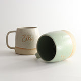 White 'Ellie' mug next to 'Paul' green mug on its side