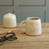 Handmade Stoneware personalised handwriting mug with 'BEST DAD' on the front. The mug sits on a wooden table next to a matching ceramic suagr pot, a copper spoon and black-rimmed glasses nearby.