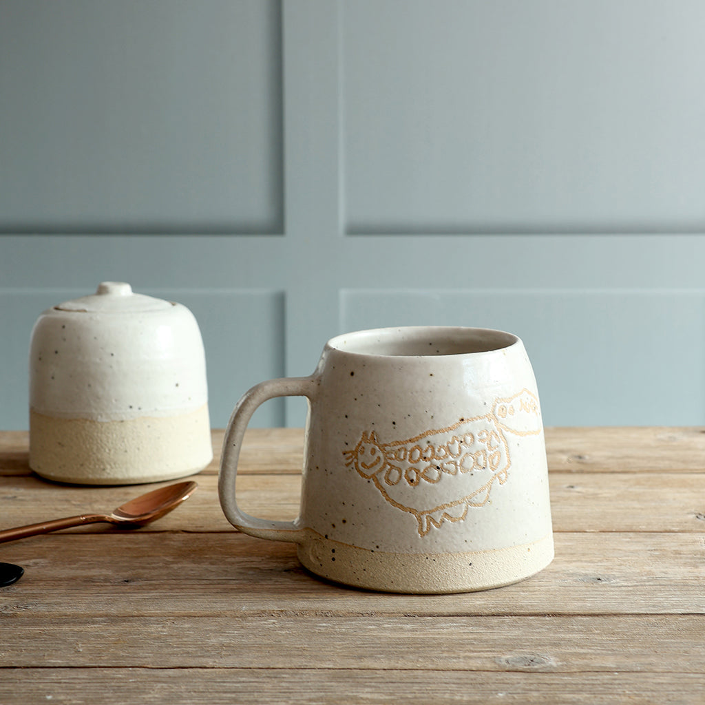 Custom Child's Drawing Stoneware mug with drawing, glazed in our off-white glaze. The mug is sat on a wooden surface with a sugar pot in the background