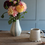 A handmade ceramic vase with a blue and white striped pattern holding a bouquet of pink, yellow, and burgundy dahlia flowers, set on a rustic wooden table next to a handmade white ceramic mug.