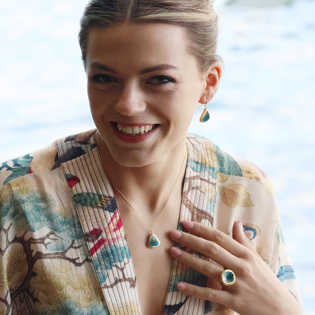 A women wearing a teardrop earring made from ceramic and teal glass with gold detailing, with a matching teardrop necklace and round ring. 
