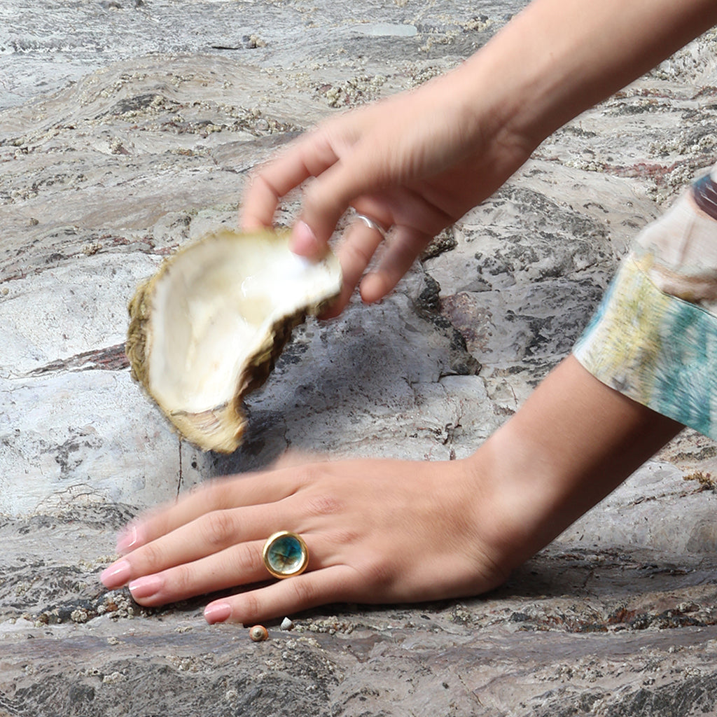 A close-up lifestyle shot of a women's hands on a rocky, sandy surface, wearing a sterling silver ring with ceramic, glass and gold charms. The glass is teal, white and brown. One hand holds a oyster shell. 