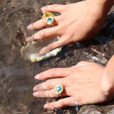 Two hands reaching into shallow water while wearing two sterling silver rings with ceramic, teal and white glass and gold round charms. 