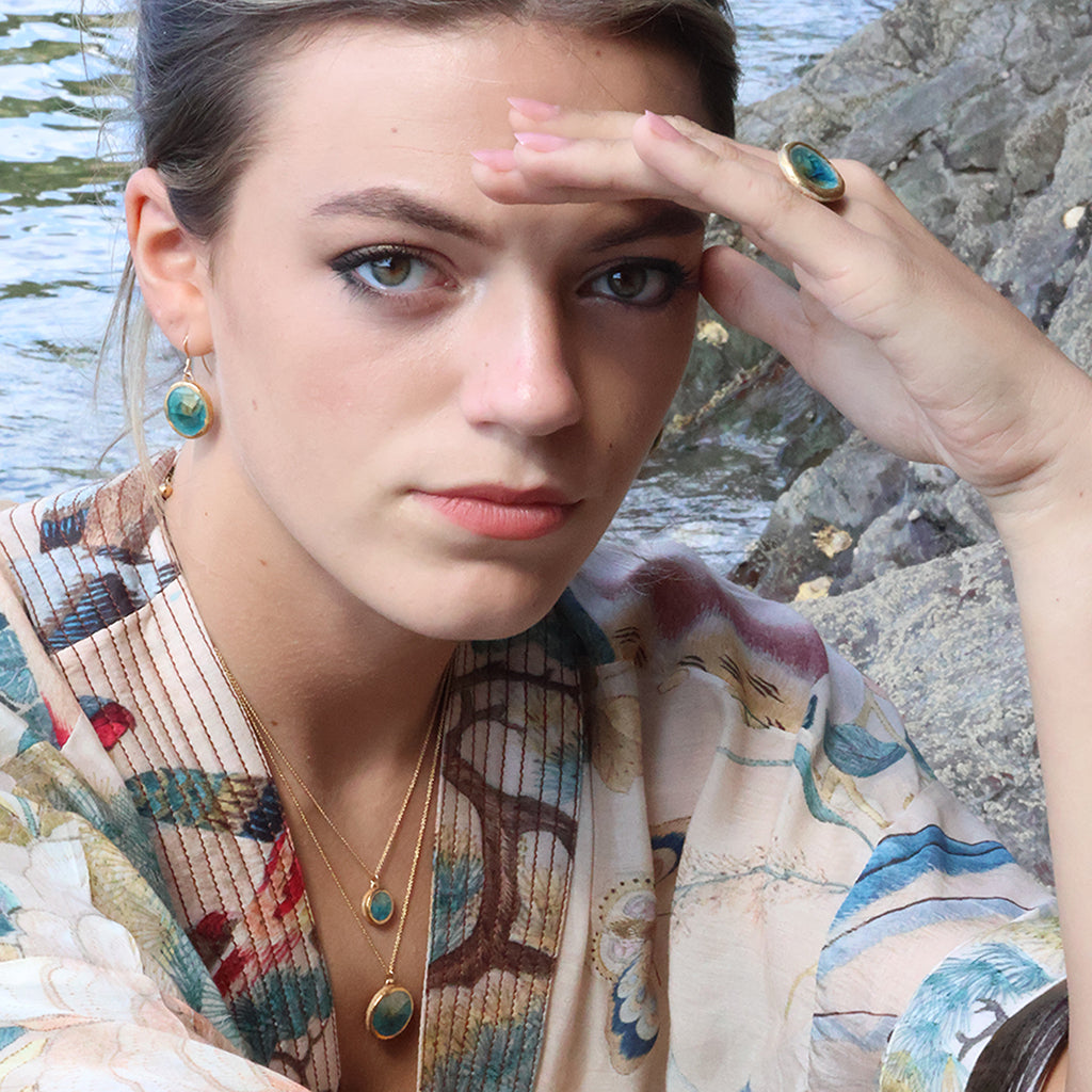 A close-up lifestyle shot of a woman wearing matching teal ceramic and glass jewellery with gold detailing, set against a rocky shoreline background. She wears a ring, two pendant necklaces and oval drop earrings. 