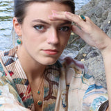 A close-up lifestyle shot of a woman wearing matching teal ceramic and glass jewellery with gold detailing, set against a rocky shoreline background. She wears a ring, two pendant necklaces and oval drop earrings. 