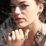 A close-up lifestyle shot of a woman wearing a gold necklace with a rectangular teal and white pendant made from ceramic and glass with gold detailing, along with matching earrings and a ring, set against a dark, natural outdoor background.