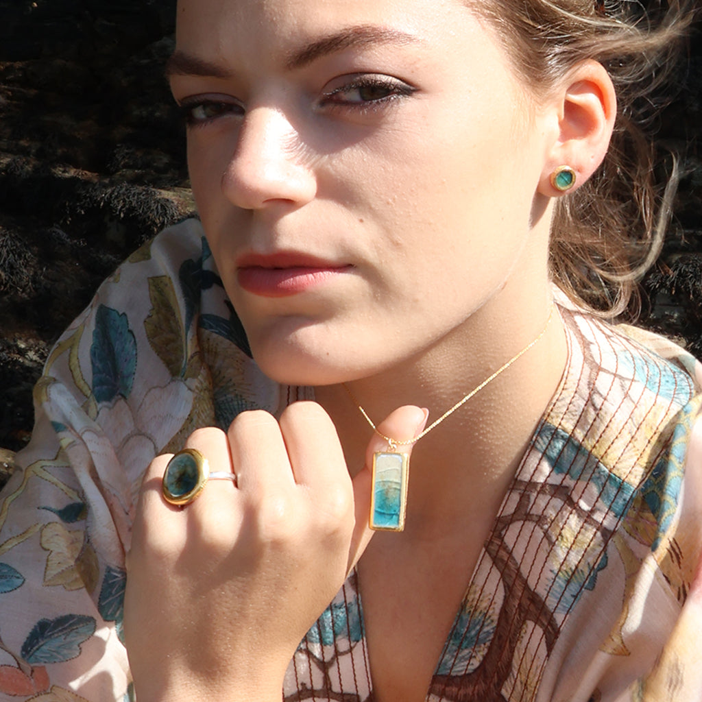 A women wears a rectangle drop pendant necklace made from ceramic and teal coloured glass, with a matching stud earring and ring. 