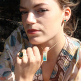 A women wears a rectangle drop pendant necklace made from ceramic and teal coloured glass, with a matching stud earring and ring. 