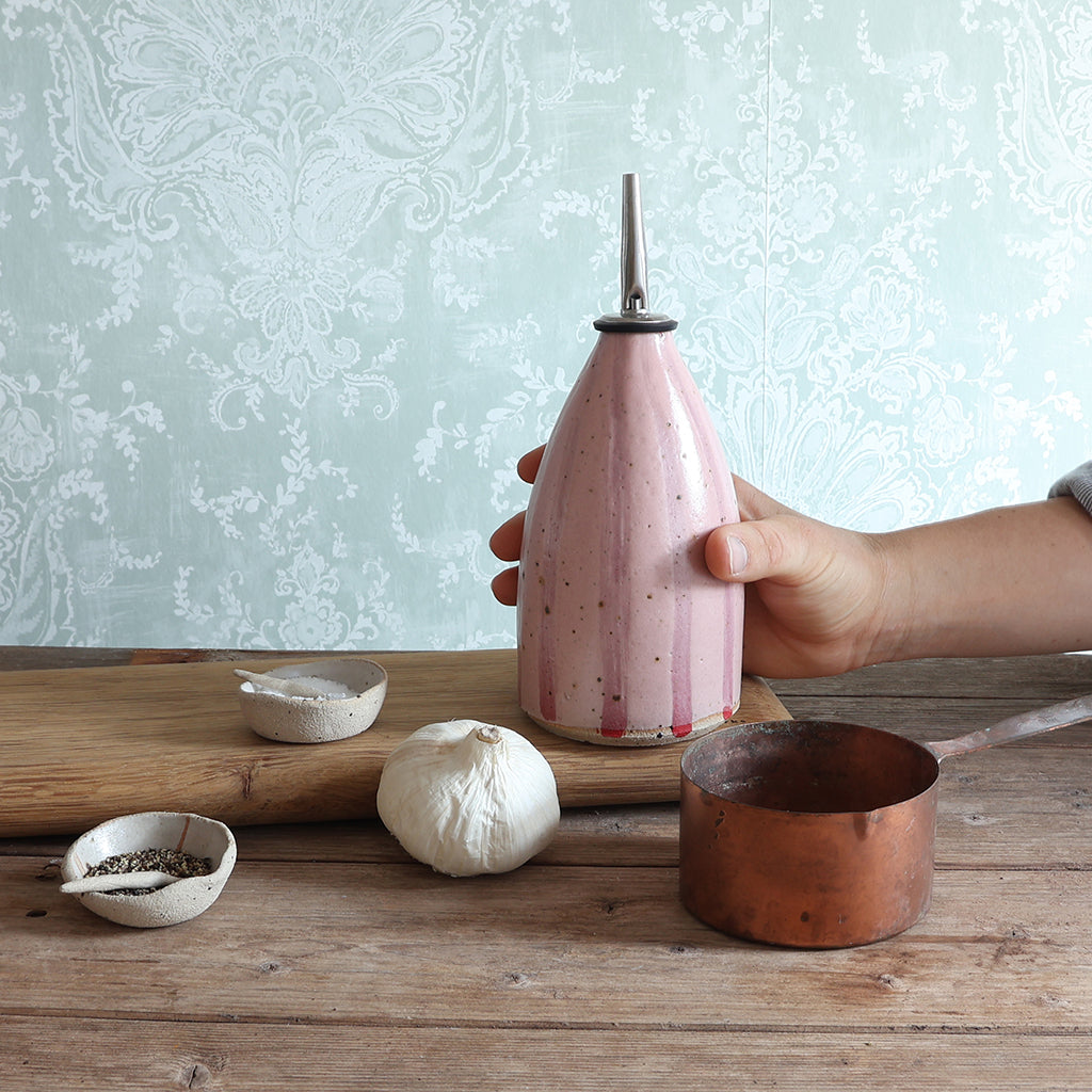 A hand holds a pink striped, flecked ceramic oil dispenser with a metal pour spout on a rustic wooden table. A copper measuring cup, a bulb of garlic, and small bowls sit nearby against a pale patterned wallpaper background.