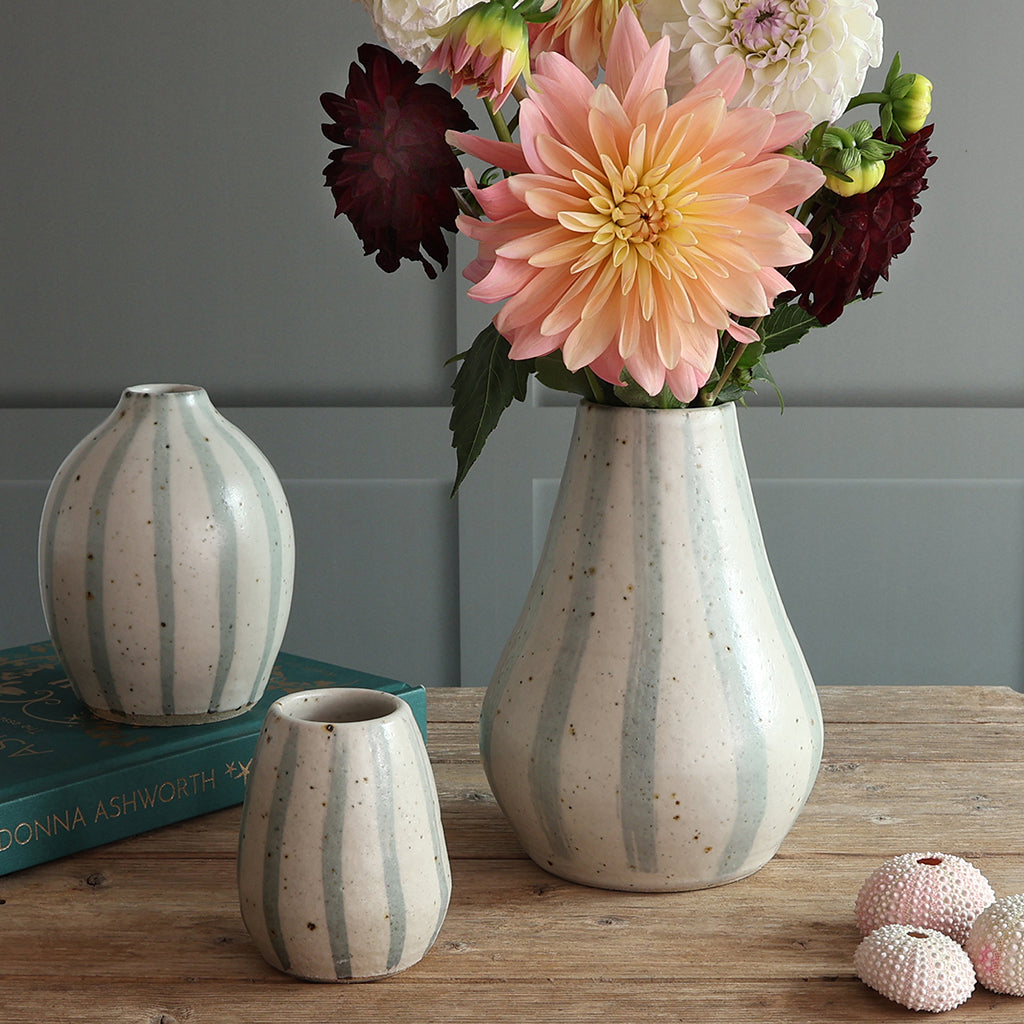 Three handmade ceramic vases with blue and white stripe designs sit on a wooden surface. The largest vase holds a bouquet of pink and burgundy dahlias on a rustic wooden table.