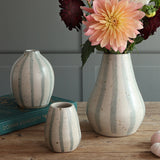 Three handmade ceramic vases in a home setting on a wooden table, with one large vase holding a peach dahlia flower, another on a teal book, and a smaller one in front. The vases feature a blue and white stripe design. 
