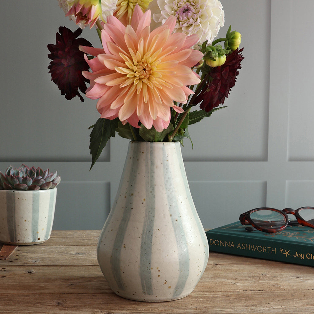 A handmade ceramic vase with vertical blue and white stripes sits on a wooden table, filled with a bouquet of dahlias including a large peach-pink flower, burgundy blooms, and white flowers. A small matching planter and a book are nearby against a paneled wall.