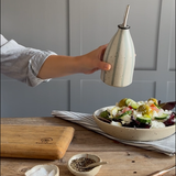 A hand holds a flecked, blue striped ceramic oil bottle with a metal pourer above a salad bowl on a wooden table, with a cutting board and small bowls in the foreground.