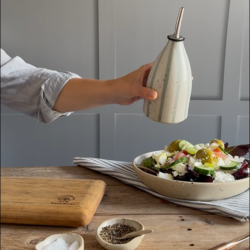 A hand holds a flecked, blue striped ceramic oil bottle with a metal pourer above a salad bowl on a wooden table, with a cutting board and small bowls in the foreground.