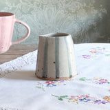 A handmade ceramic jug with a tapered shape, featuring vertical light blue stripes on a speckled off-white glaze, set on a white embroidered tablecloth next to a pink mug.