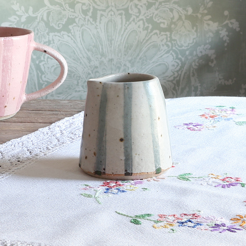 A handmade ceramic jug with a tapered shape, featuring vertical light blue stripes on a speckled off-white glaze, set on a white embroidered tablecloth next to a pink mug.
