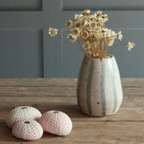 A handmade ceramic vase with blue and white stripes, filled with dried flowers, sits on a rustic wooden table. Three pale pink sea urchin shells are placed in front of it against a gray paneled wall background. 