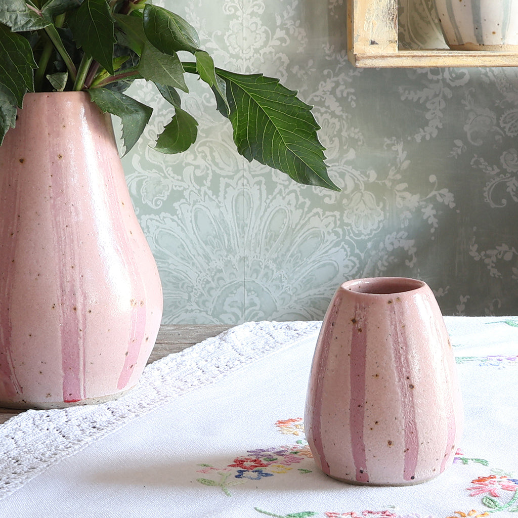 Two pink stripey handmade ceramic vases on a white lace vintage tablecloth, with one vase holding green leaves against a patterned wallpaper background.