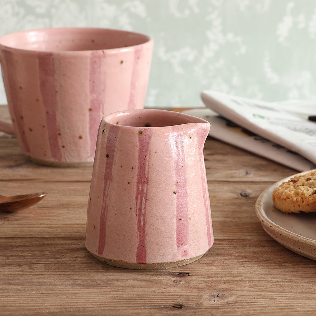 A pink stripy speckled mini ceramic jug with a matching mug in the background, styled on a rustic wooden table with a plate of food and a newspaper.