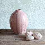 A handmade rounded ceramic vase with a pink stipe design stands on a rustic wooden table, next to three pink sea urchin shells. 