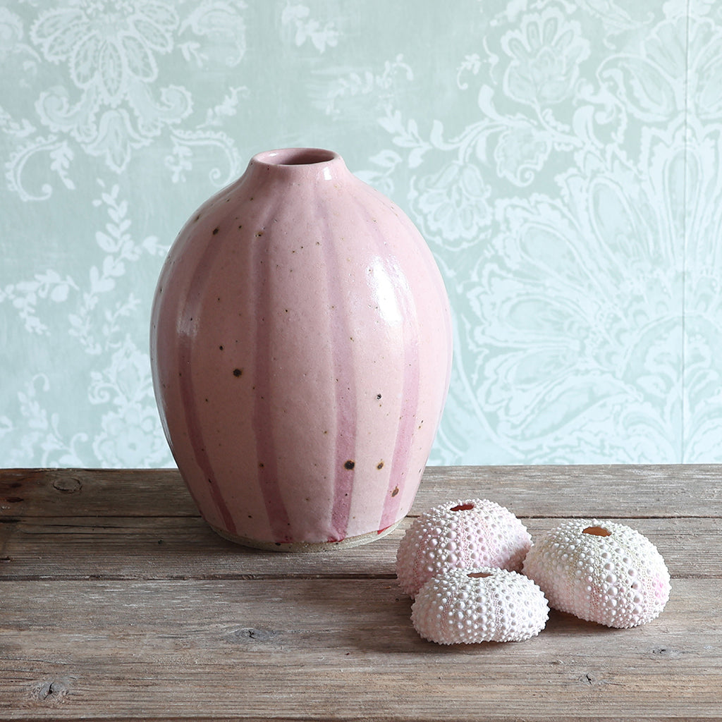 A handmade rounded ceramic vase with a pink stipe design stands on a rustic wooden table, next to three pink sea urchin shells. 