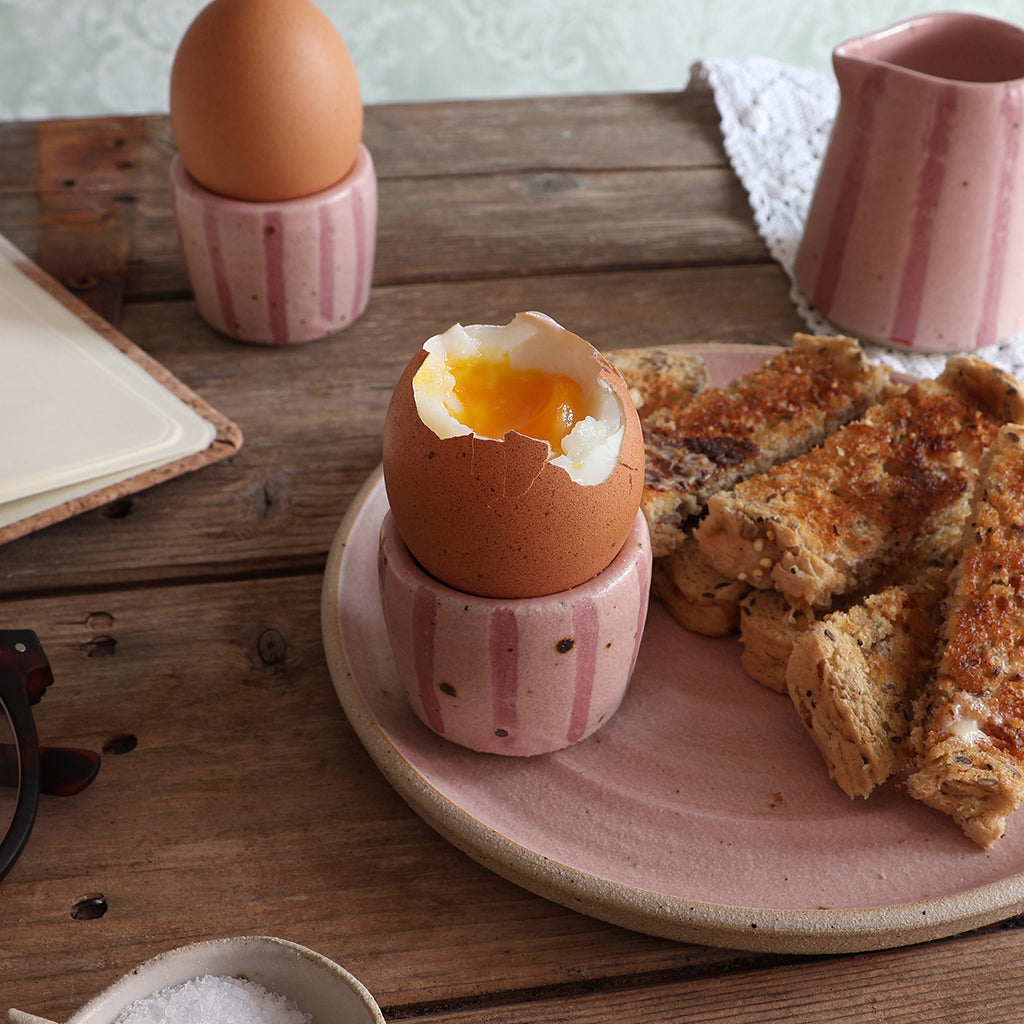 A breakfast scene on a rustic wooden table featuring a soft-boiled egg in a handmade pink striped ceramic egg cup, alongside toast on a pink plate, another egg in a matching egg cup, and a matching small jug. 