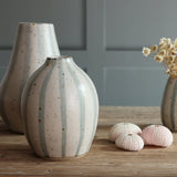 Three handmade ceramic vases with blue and white stripe designs arranged on a wooden table, with a collection of three pink sea urchin shells. 