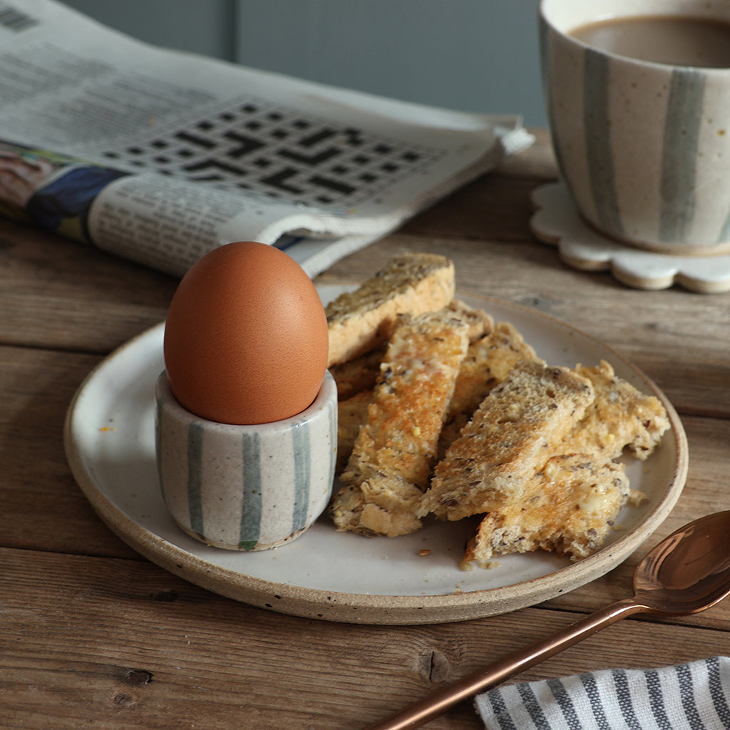 A ceramic breakfast scene on a wooden table with a handmade white plate holding a brown egg in a blue and white striped egg cup and several pieces of toast. A matching mug and brass spoon are visible in the background, along with a newspaper.