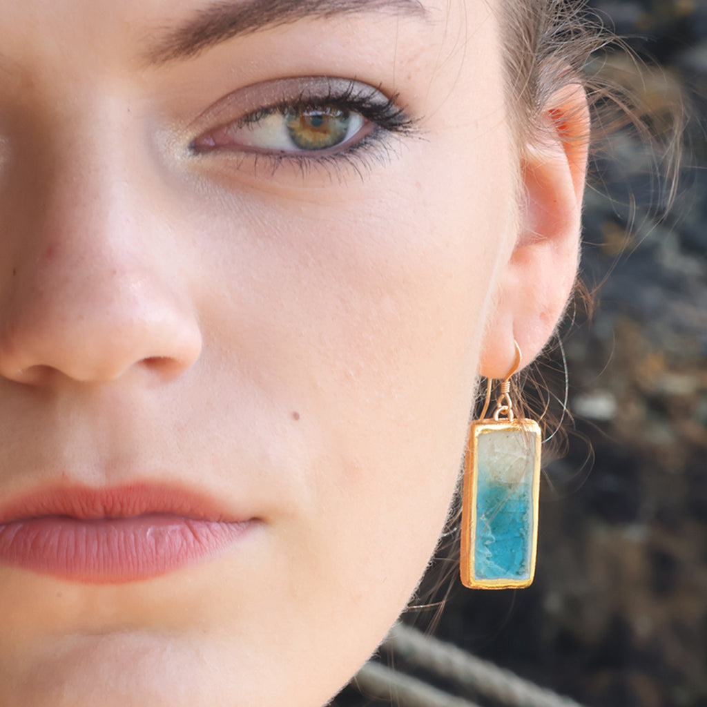 Close-up of a woman wearing a ceramic drop earring with teal and white glass and gold detailing. 