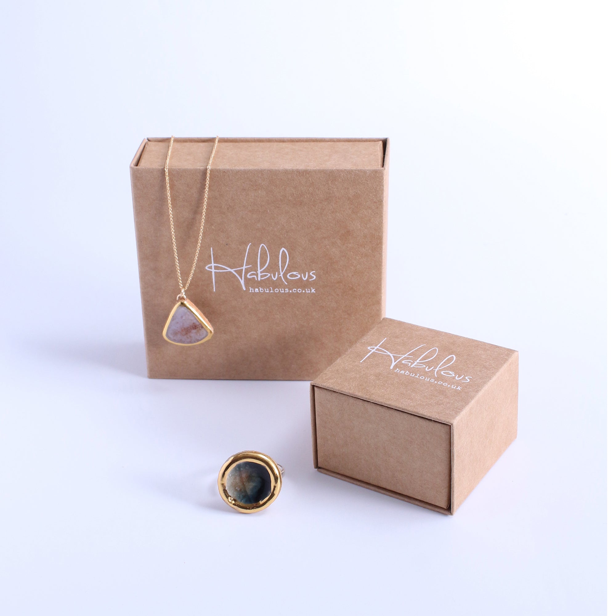 Cardboard jewellery packaging with Habulous branding pictured against a white background. Our Dusk Teardrop necklace with a gold chain and ceramic, glass and gold lustre pendant hangs over the box, and our large Eclipse Ring sits in the foreground. 