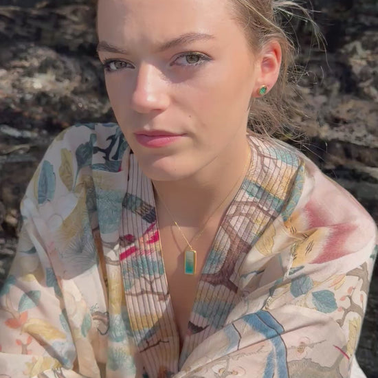 A women wears gold and teal ceramic and glass stud earrings, with a matching rectangular pendant necklace and rings, set against a rocky background. 