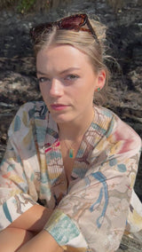 A women wears gold and teal ceramic and glass stud earrings, with a matching rectangular pendant necklace and rings, set against a rocky background. 