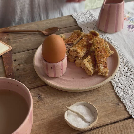 A person uses a brass spoon to crack open a boiled egg sat in a handmade pink stripe egg cup sat on a pink plate filled with toast. A matching mug and small  jug also sit on the wooden table. 
