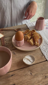 A person uses a brass spoon to crack open a boiled egg sat in a handmade pink stripe egg cup sat on a pink plate filled with toast. A matching mug and small  jug also sit on the wooden table. 