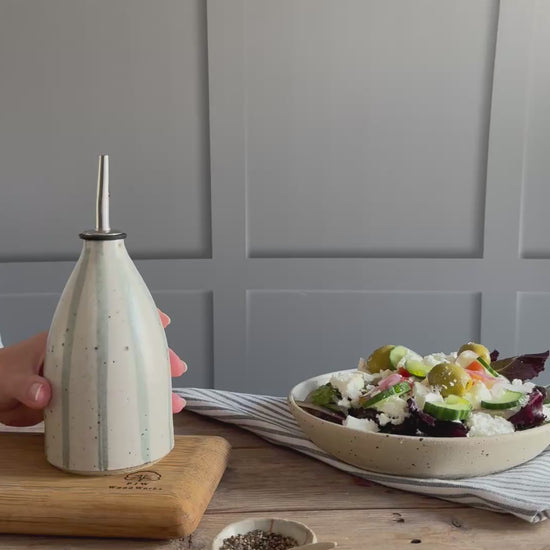 A person picks up a handmade ceramic oil pourer with a blue and white stripe design and pours oil from the bottle onto a salad in a handmade bowl. 