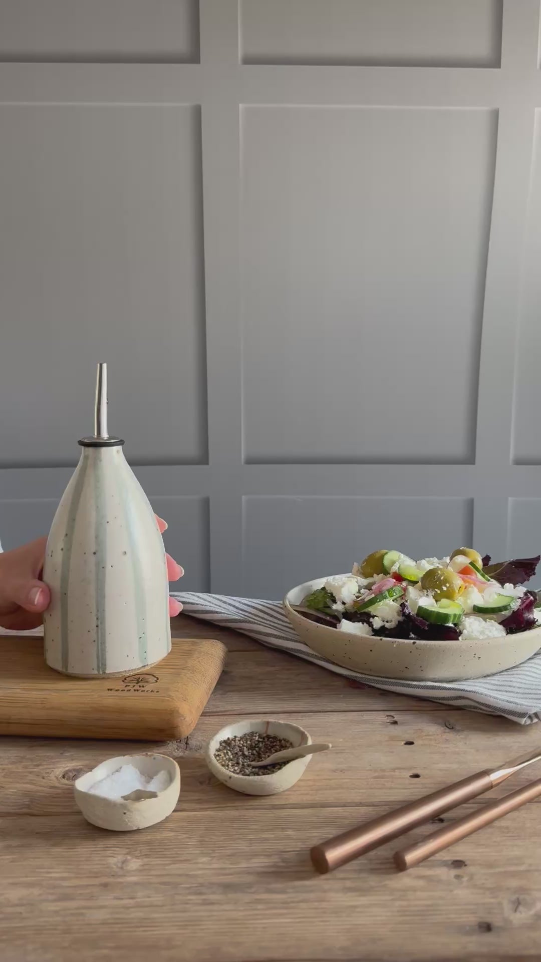 A person picks up a handmade ceramic oil pourer with a blue and white stripe design and pours oil from the bottle onto a salad in a handmade bowl. 