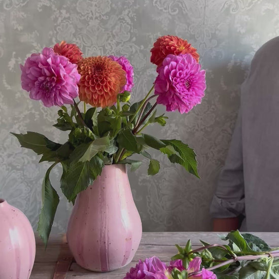 Three handmade ceramic vases with pink striped designs arranged on a wooden surface. A person places a flower into the bouquet inside the largest vase.
