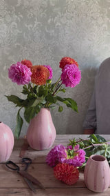 Three handmade ceramic vases with pink striped designs arranged on a wooden surface. A person places a flower into the bouquet inside the largest vase.