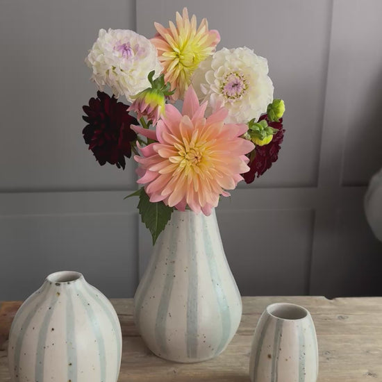 Three handmade ceramic vases with blue and white striped designs arranged on a wooden surface. A person places a flower into the bouquet inside the largest vase. 
