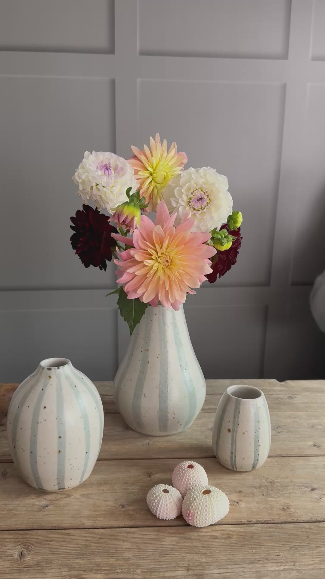 Three handmade ceramic vases with blue and white striped designs arranged on a wooden surface. A person places a flower into the bouquet inside the largest vase. 